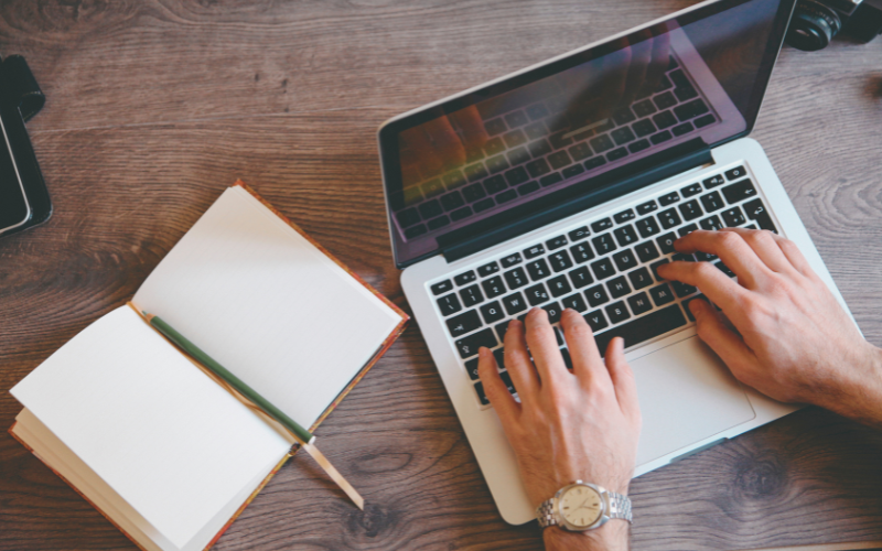 man typing on silver laptop with open note book and pencil